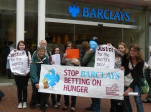 A group of people hold a banner in front of a Barclays bank that reads 'Stop Barclays Betting on Hunger'.
